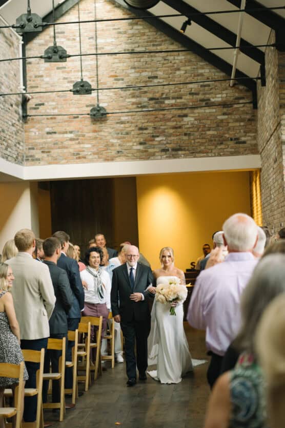 A bride in a white dress holding a bouquet walks down the aisle with an older man, likely her father, in a venue with brick walls, while guests stand and watch. by Justin Salem Meyer