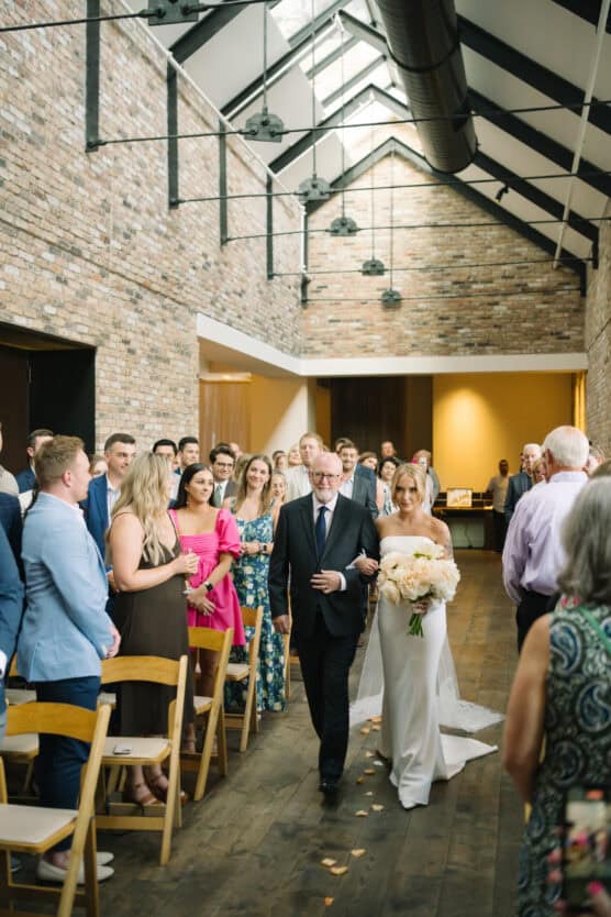 A bride in a white dress walks down the aisle holding a bouquet, accompanied by an older man in a suit. Guests on both sides of the aisle watch and smile in a bright, modern venue with brick walls and wooden floors. by Justin Salem Meyer