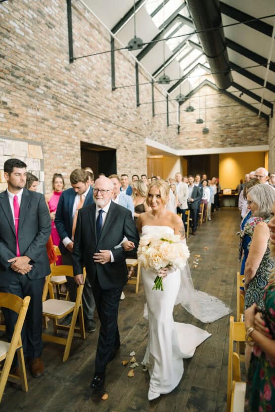 A bride in a white dress holding a bouquet walks down the aisle with an older man in a suit. Guests seated on both sides watch as they make their way through a sunlit, modern venue with brick walls and wooden floors. by Justin Salem Meyer