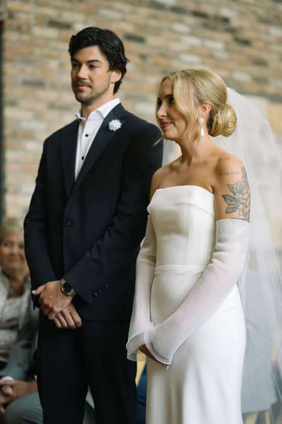 A bride in an off-shoulder white dress with sheer sleeves and a veil stands beside a groom in a black suit and white shirt. They face forward during a wedding ceremony, with guests seated behind them. by Justin Salem Meyer