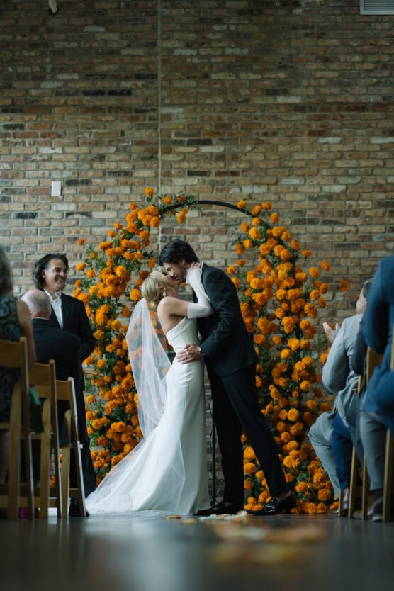 A bride and groom share a kiss at their wedding ceremony, standing in front of a circular floral arch of orange flowers, with guests seated on either side and a brick wall in the background. by Justin Salem Meyer