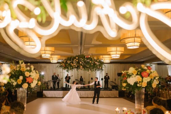 A bride and groom dance together in an elegant ballroom with a live band on stage, floral arrangements, and warm lighting. Large illuminated letters are visible at the top of the image. by Justin Salem Meyer