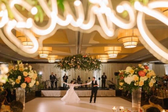 A bride and groom share their first dance in a grand ballroom decorated with large floral arrangements, hanging greenery, and soft, glowing lights while a live band plays on stage in the background. by Justin Salem Meyer