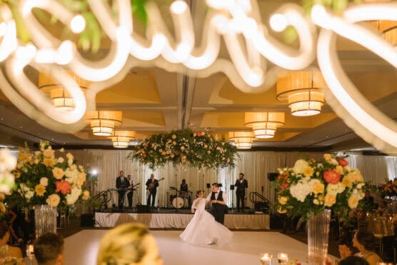 A bride and groom share their first dance in an elegant ballroom, surrounded by large flower arrangements and warm lighting, while a live band performs in the background. A glowing sign is partially visible at the top. by Justin Salem Meyer