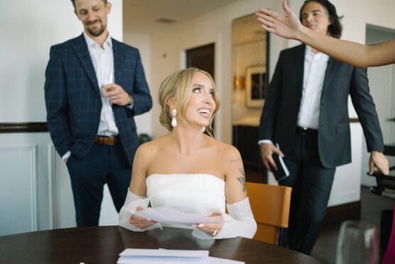 A smiling woman in an off-the-shoulder white dress sits at a table holding papers, while two men in suits stand behind her. An outstretched hand enters from the right. The setting appears to be an indoor office or formal room. by Justin Salem Meyer