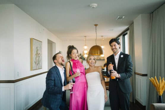 Four people dressed formally celebrate indoors; one man in a tuxedo opens a bottle of champagne as the others, holding glasses, react with excitement and joy. The setting appears modern and elegant with warm lighting. by Justin Salem Meyer