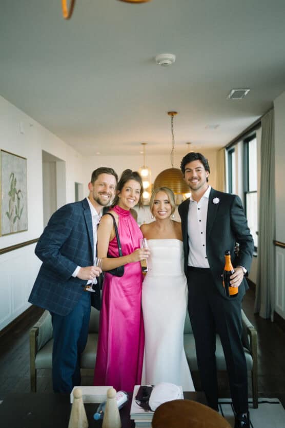 Four people pose and smile indoors, dressed formally; two men in suits and two women, one in a bright pink dress and the other in a strapless white dress, stand together holding drinks in a modern, well-lit room. by Justin Salem Meyer