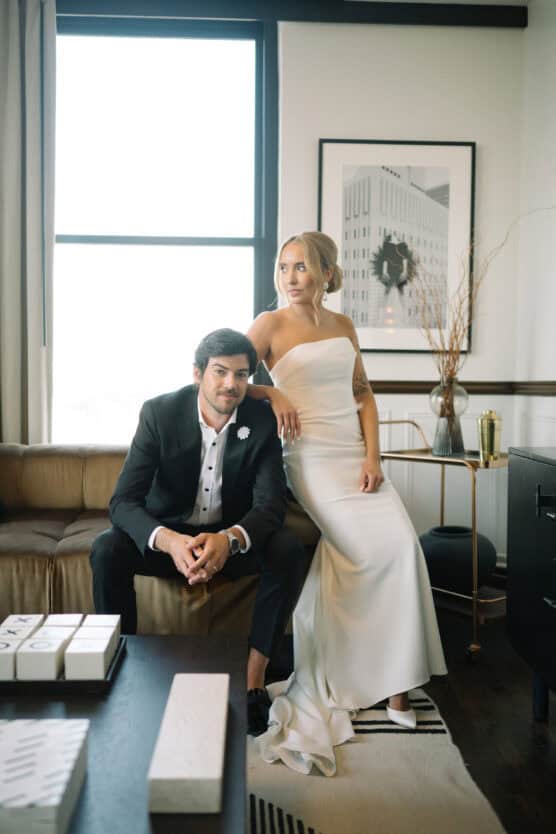 A bride in a white one-shoulder gown stands beside a groom in a black suit seated on a brown couch, both posing stylishly in a modern, sunlit living room. by Justin Salem Meyer