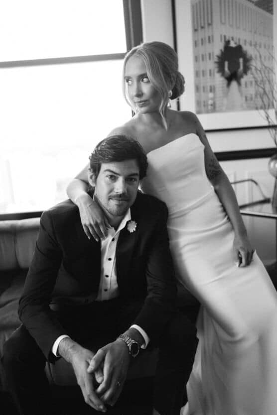 A black and white photo of a bride in a strapless gown and a groom in a suit. The bride stands next to the seated groom, resting her hand on his shoulder. They pose in a modern room with large windows and framed art. by Justin Salem Meyer