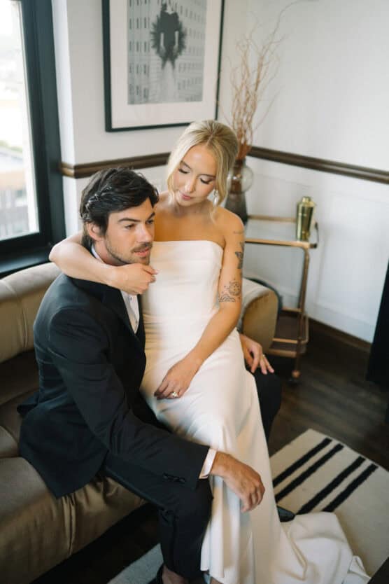 A bride in a strapless white dress sits on a couch beside a groom in a black suit, her arm around his shoulders. They appear calm and intimate in a modern, softly lit room with framed artwork and decor. by Justin Salem Meyer