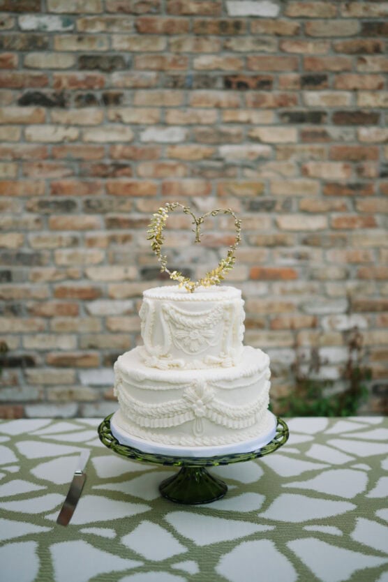 A two-tier white wedding cake with ornate icing details and a gold heart topper sits on a green cake stand atop a patterned tablecloth, with a brick wall in the background. A cake knife is placed beside the cake. by Justin Salem Meyer