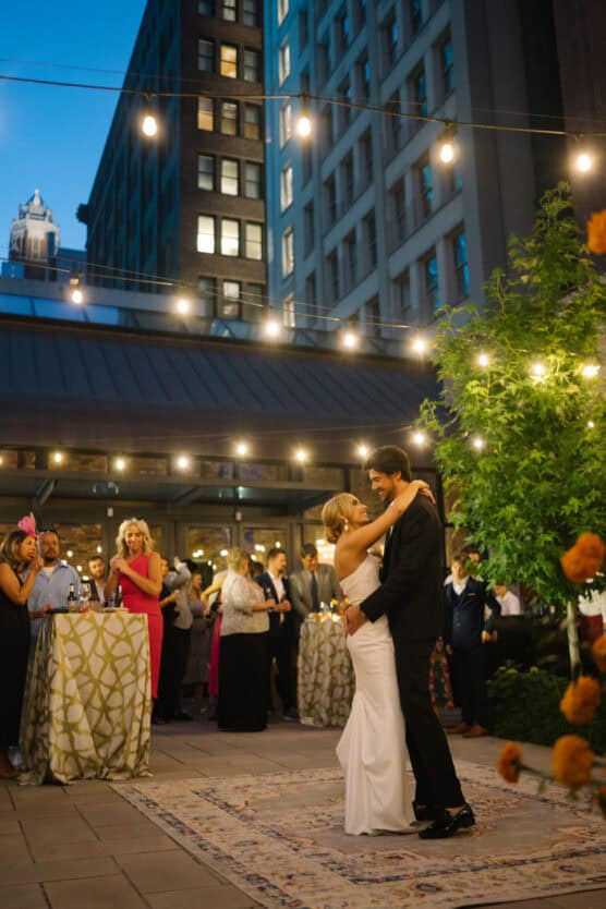 A bride and groom share a first dance on an outdoor rooftop, surrounded by guests and string lights, with tall buildings in the background during the evening. by Justin Salem Meyer