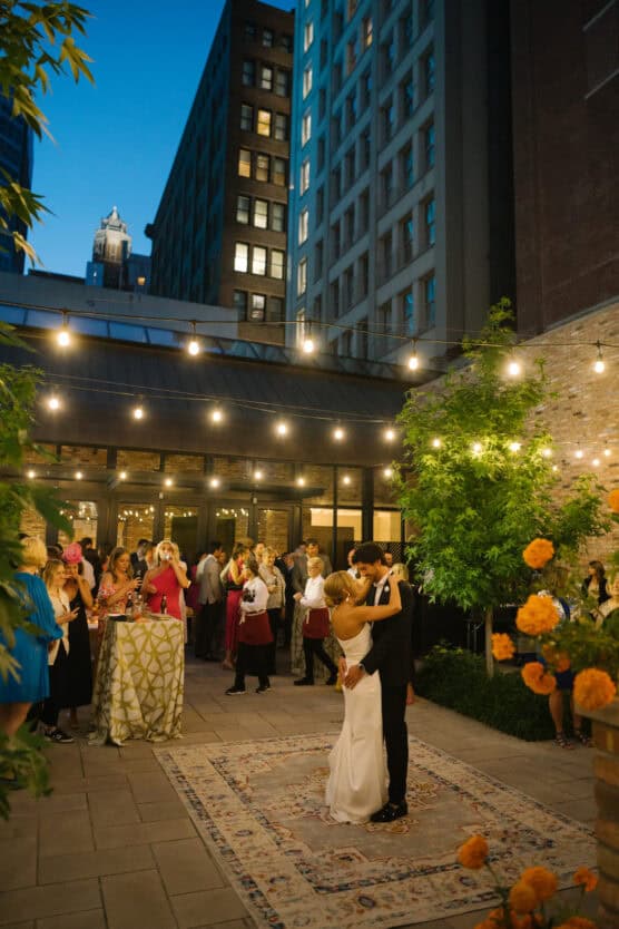 A couple shares a first dance on a rug at an outdoor evening wedding reception, surrounded by guests and string lights, with tall city buildings in the background and vibrant orange flowers in the foreground. by Justin Salem Meyer