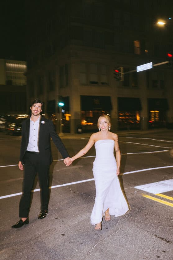 A smiling couple dressed in wedding attire hold hands and walk across a city street at night, illuminated by streetlights and surrounded by tall buildings. by Justin Salem Meyer