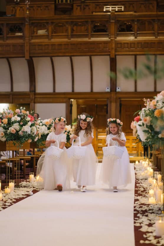 Three young flower girls in white dresses and floral crowns walk down a candle-lit aisle in a decorated church, each carrying a white basket. The aisle is lined with flower arrangements and rose petals. by Justin Salem Meyer