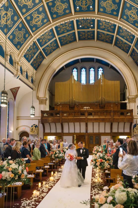 A bride in a white gown walks down a flower-lined aisle with her father in a grand church, as guests stand and look on. The church has a blue vaulted ceiling, stained glass windows, and a large golden pipe organ. by Justin Salem Meyer