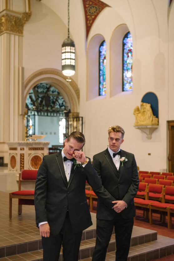 Two men in tuxedos stand in a church. One wipes away a tear, while the other stands supportively beside him. Stained glass windows and red chairs are visible in the background. by Justin Salem Meyer