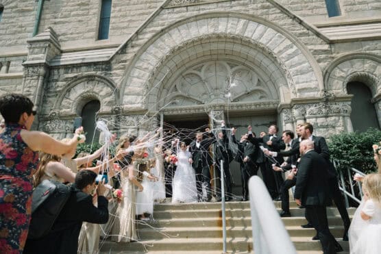 A bride and groom exit a stone church as guests on both sides spray them with white streamers. The bridal party stands around, smiling and celebrating on the church steps. by Justin Salem Meyer