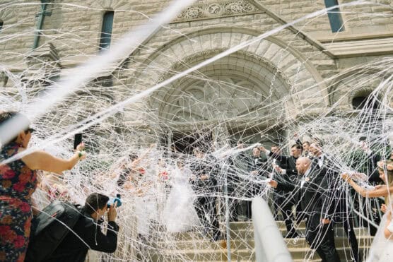 A wedding party exits a stone church while guests spray streams of white silly string, filling the air with tangled ribbons as people celebrate on the steps, some taking photos. by Justin Salem Meyer