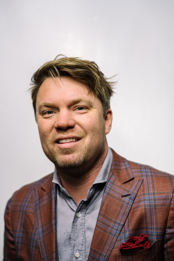 A man with blond hair wearing a plaid brown blazer over a light blue shirt smiles in front of a plain white background. by Justin Salem Meyer