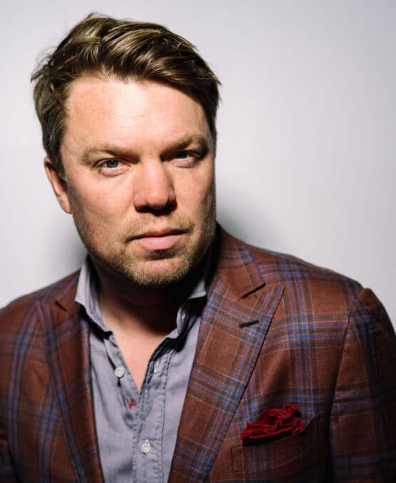 A man with light skin and short, styled brown hair wears a brown and blue plaid blazer over a gray shirt. He stands against a plain white background, looking directly at the camera with a neutral expression. by Justin Salem Meyer