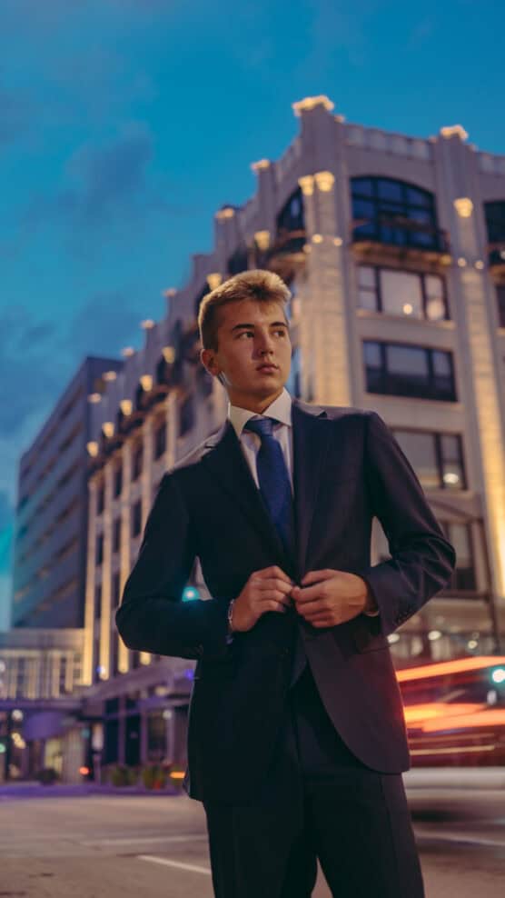 A young man in a suit stands confidently on a city street at dusk, with a lit-up building and blurred car lights in the background. by Justin Salem Meyer