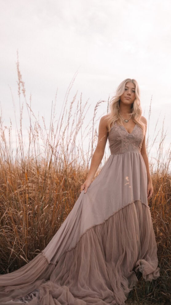 A woman with long blonde hair in a flowing taupe dress stands in a field of tall, dry grass under a cloudy sky, looking calmly at the camera. by Justin Salem Meyer