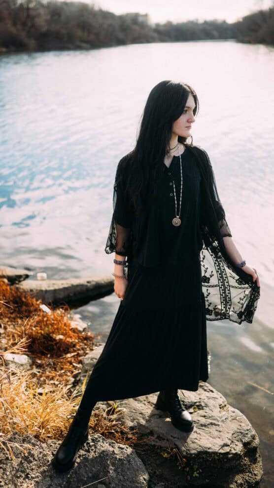 A woman in a black dress and boots stands on a rock by a river, looking to the side. She wears layered necklaces and a lace shawl, with autumn foliage and water in the background. by Justin Salem Meyer
