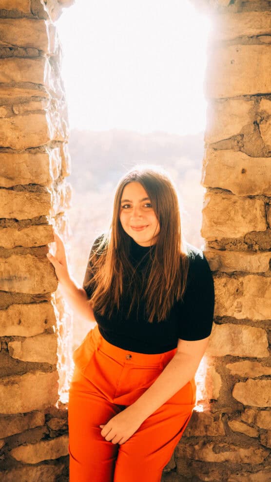 A woman with long brown hair, wearing a black top and bright orange pants, sits in a stone window frame with sunlight shining behind her, creating a warm, glowing atmosphere. by Justin Salem Meyer