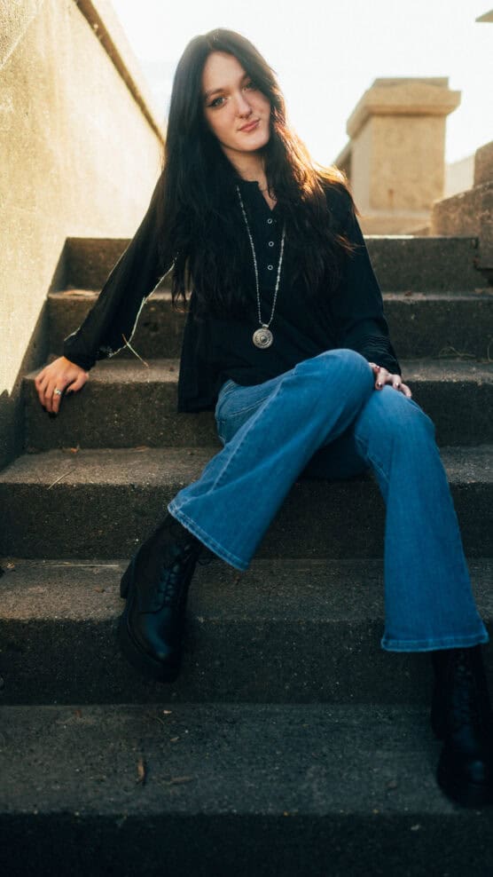 A woman with long dark hair sits on outdoor concrete steps, wearing a black top, blue jeans, black boots, and a pendant necklace. She is smiling and posing casually in natural sunlight. by Justin Salem Meyer