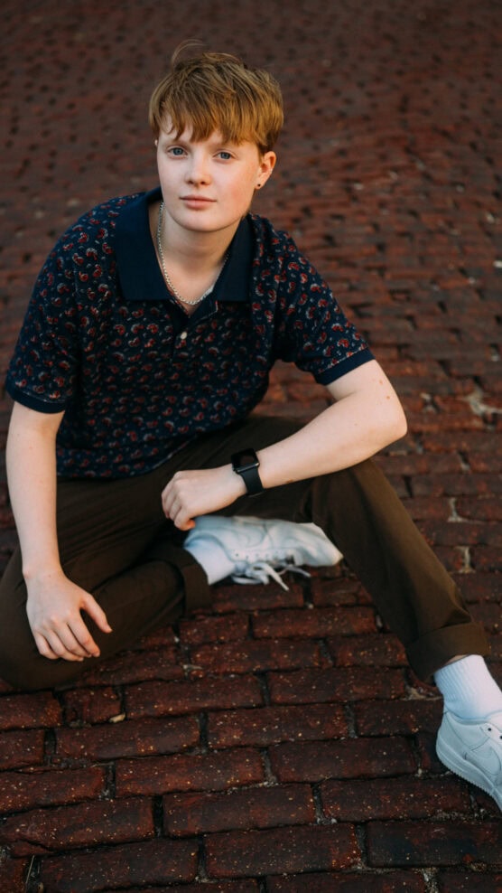 A person with short, light brown hair sits on a red brick pavement. They wear a navy patterned polo shirt, brown pants, white sneakers, a silver chain necklace, and a smartwatch, looking calmly at the camera. by Justin Salem Meyer