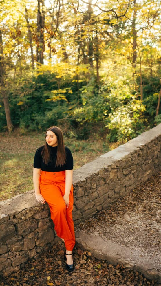 A woman with long brown hair, wearing a black top and bright orange pants, sits on a stone wall in a park with trees and fallen leaves. Sunlight filters through the autumn foliage in the background. by Justin Salem Meyer