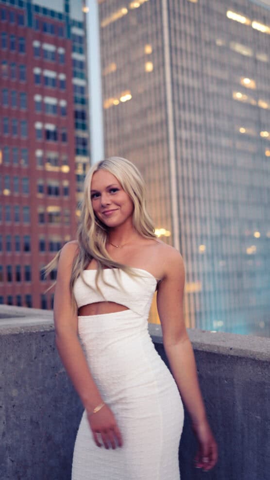 A woman in a fitted white dress stands on a rooftop at dusk, smiling, with tall city buildings in the background. by Justin Salem Meyer