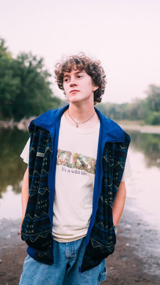 A young person with curly hair stands near a calm river, wearing a patterned blue vest over a white t-shirt that says Its a wild life. Trees and greenery are visible in the background. by Justin Salem Meyer