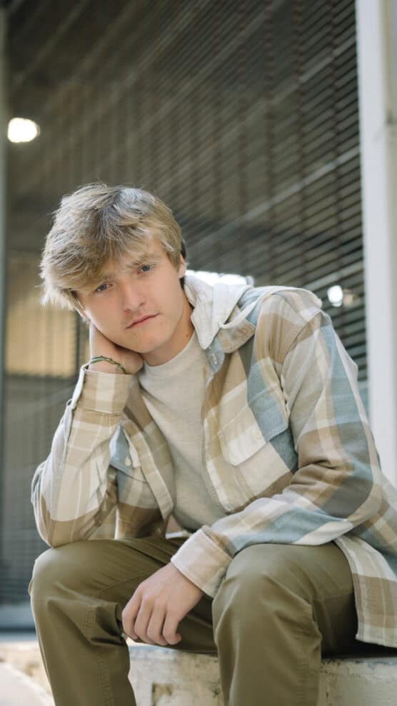 A young person with blonde hair, wearing a plaid shirt over a gray t-shirt and khaki pants, sits with their head resting on one hand, looking thoughtfully at the camera. A metal fence is in the background. by Justin Salem Meyer
