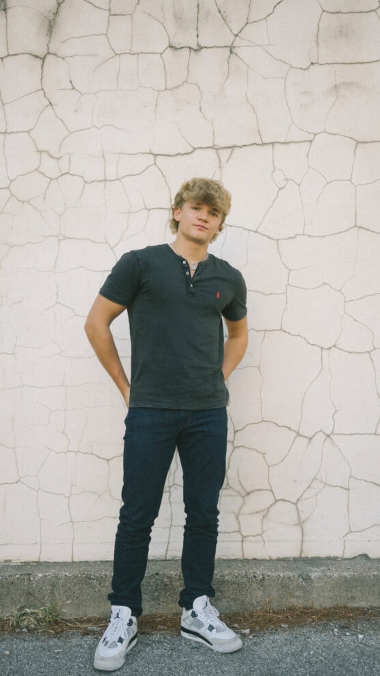 A young man with blond hair stands in front of a cracked, textured white wall, wearing a black polo shirt, dark jeans, and white sneakers. His arms are relaxed behind his back and he looks at the camera. by Justin Salem Meyer