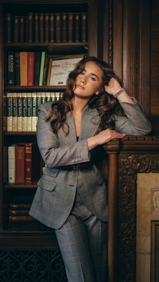 A woman in a gray plaid suit leans against a carved wooden mantle in a library, with shelves of books in the background. She has long, wavy hair and a relaxed, confident expression. by Justin Salem Meyer