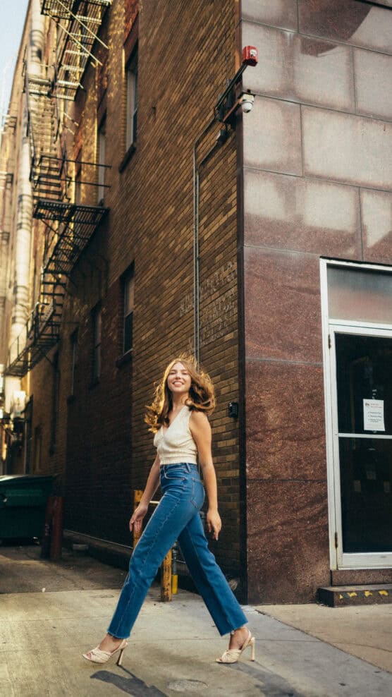 A woman with long hair, wearing a sleeveless top, blue jeans, and heeled sandals, smiles as she walks confidently down a city alley next to a brick building. by Justin Salem Meyer