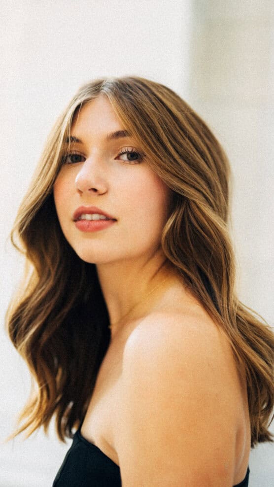 A young woman with wavy, light brown hair and bare shoulders gazes confidently at the camera against a softly blurred, light background. She is wearing a strapless black top. by Justin Salem Meyer