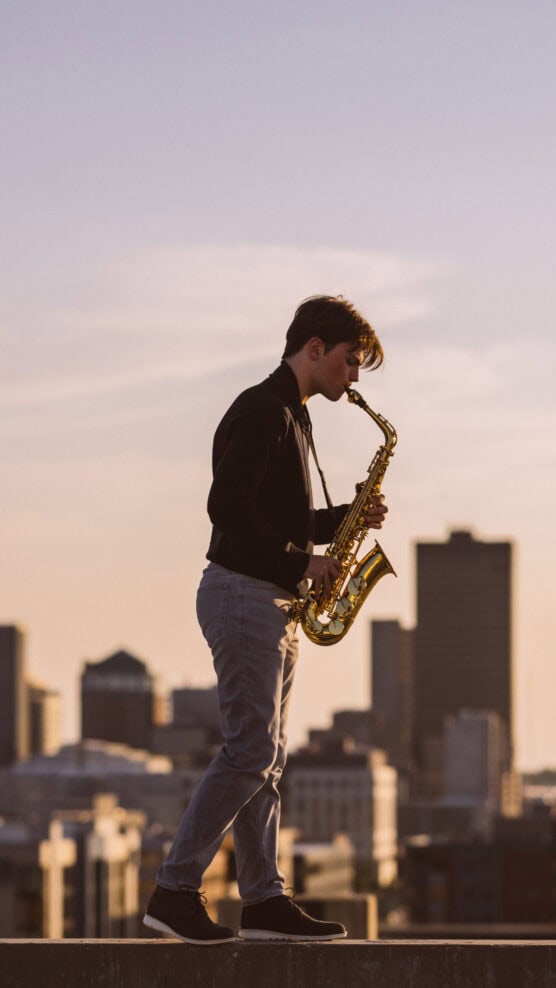 A person playing a saxophone stands on a rooftop at sunset, with a city skyline and tall buildings in the background. by Justin Salem Meyer