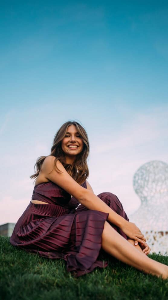 A woman in a sleeveless maroon dress sits on grass, smiling at the camera, with a clear blue sky and a white abstract sculpture in the background. by Justin Salem Meyer