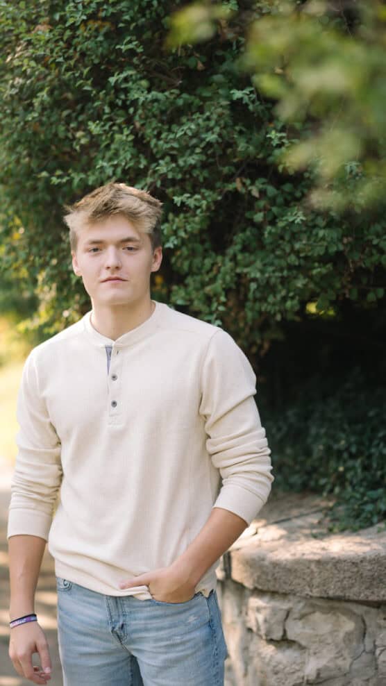 A young man with short blonde hair wearing a light-colored long-sleeve shirt and blue jeans stands outdoors near a stone wall and green bushes, looking at the camera with a neutral expression. by Justin Salem Meyer