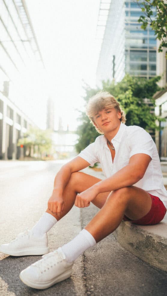 A young man with blond hair sits on a curb in a city street, wearing a white shirt, red shorts, white socks, and white sneakers. The background shows modern buildings and trees in bright sunlight. by Justin Salem Meyer