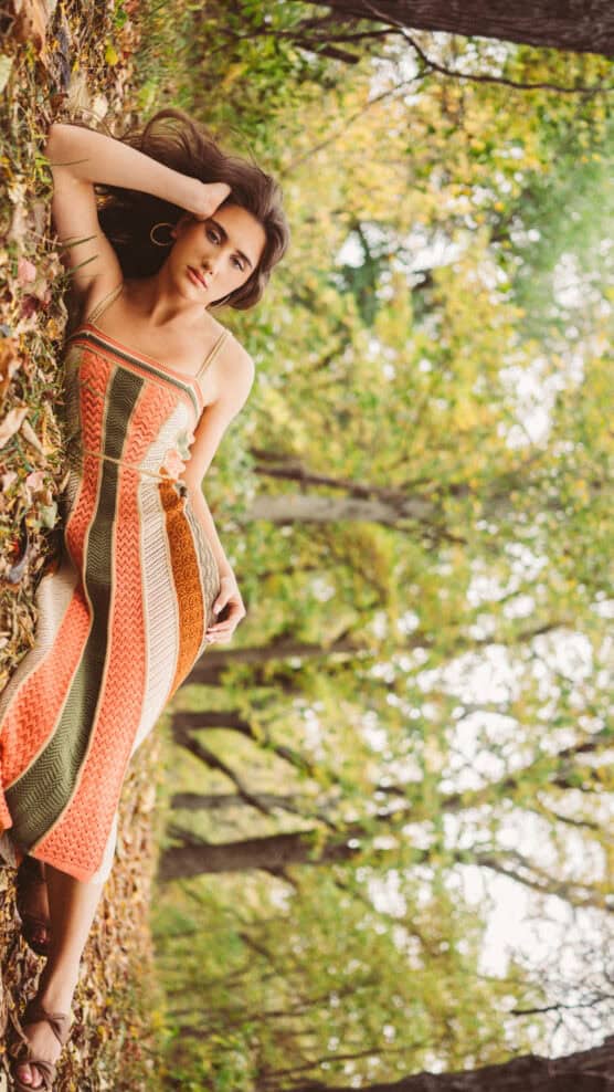 A woman in a colorful striped dress lies on the ground covered with autumn leaves in a forest, resting her head on one hand and looking at the camera. Trees with green and yellow foliage surround her. by Justin Salem Meyer