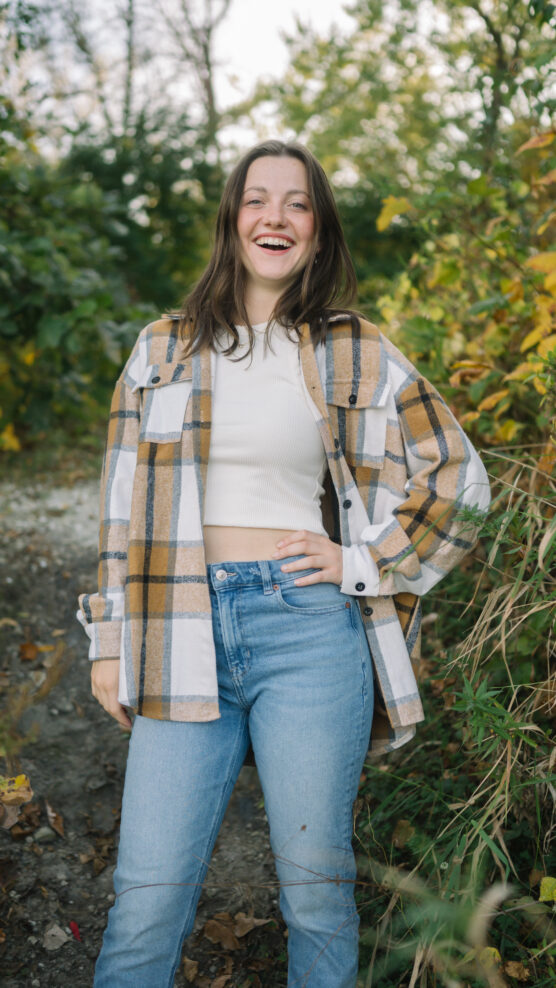 A young woman with long brown hair stands outdoors on a leafy path, smiling. She wears a white turtleneck, blue jeans, and a plaid flannel shirt. Green and yellow foliage surrounds her in the background. by Justin Salem Meyer