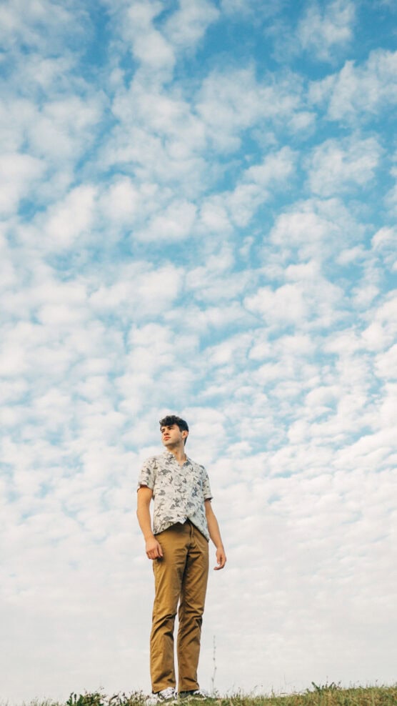 A man wearing sunglasses, a floral short-sleeve shirt, and tan pants stands on grass, looking to the side under a bright blue sky filled with scattered clouds. by Justin Salem Meyer