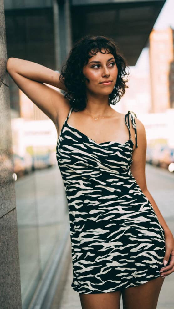 A woman with curly dark hair in a black and white zebra-print dress leans against a wall outdoors, looking to the side, with an urban street background. by Justin Salem Meyer