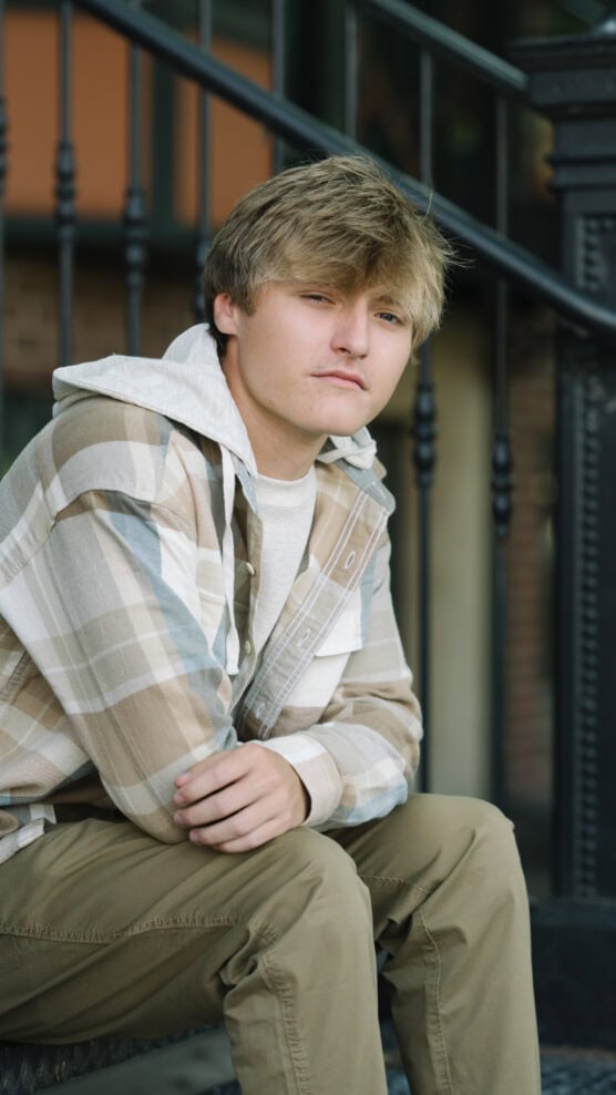 A young person with blond, tousled hair sits outdoors on steps, wearing a plaid shirt over a hoodie and khaki pants, looking at the camera with a neutral expression. by Justin Salem Meyer