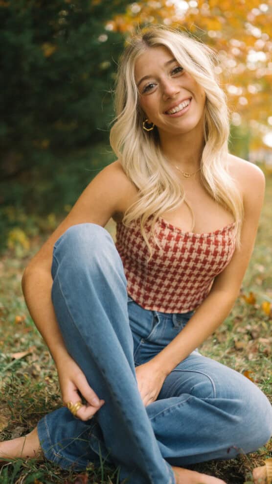A young woman with long blonde hair sits on grass outdoors, smiling. She wears a red checkered top and blue jeans, with autumn foliage in the background. by Justin Salem Meyer