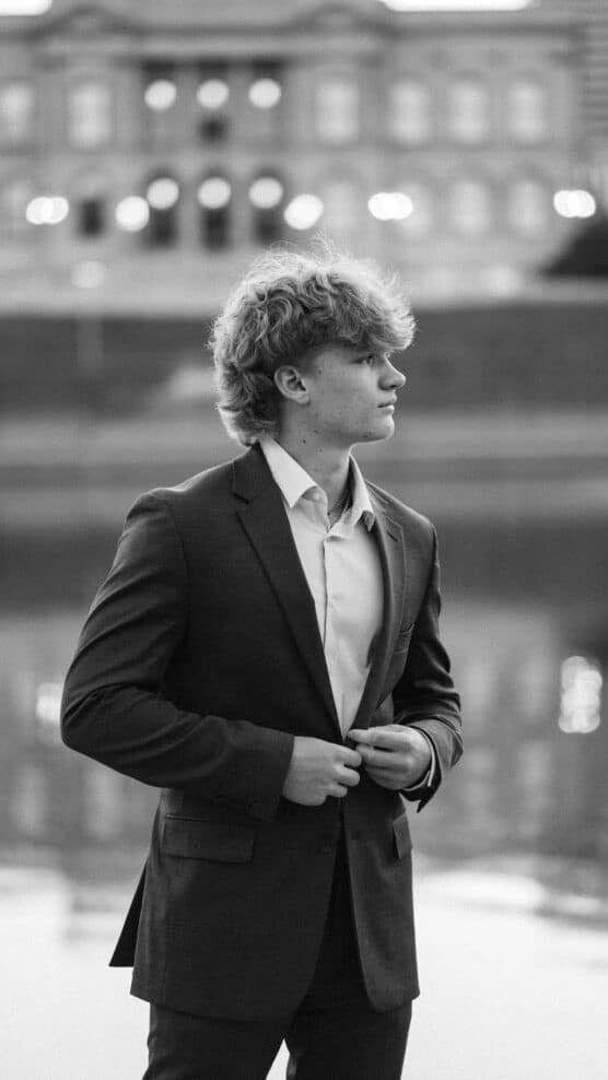 A young man in a suit stands by a body of water, buttoning his jacket and looking to the side. The background features a large, blurred building with lights. The photo is in black and white. by Justin Salem Meyer